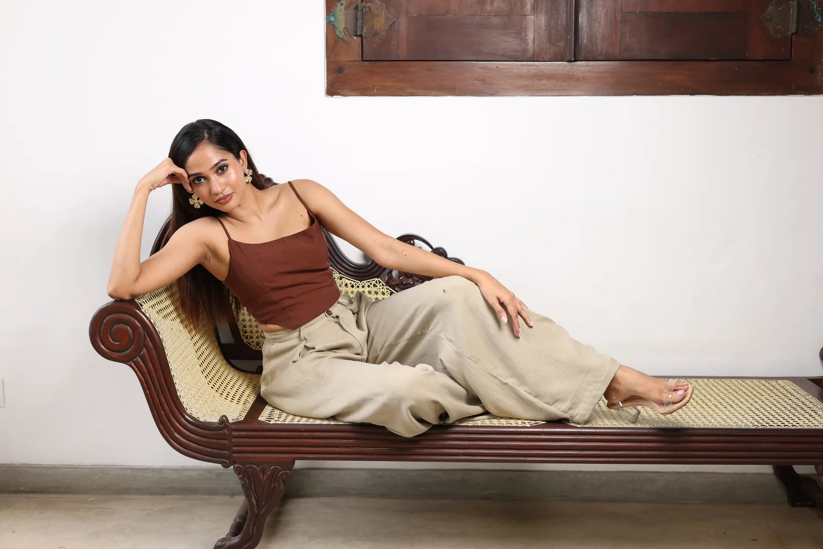 Woman in a brown camisole and wide-leg beige trousers reclining on a cane bench